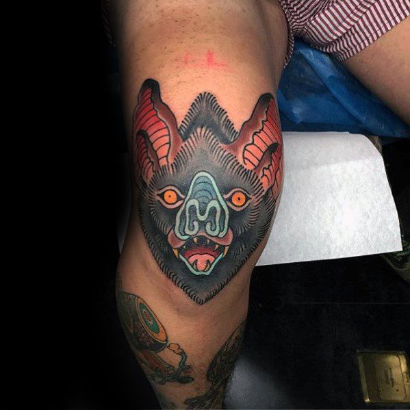 Neo-traditional bat tattoo on the thigh with bold lines, vivid red and blue tones, and fierce expression with glowing orange eyes.