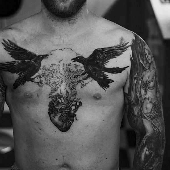 Odin’s ravens tattoo design with Huginn and Muninn flying over a detailed heart and tree of life, inked across the chest.