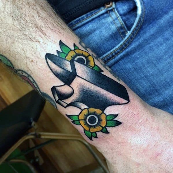 Anvil tattoo on forearm with bold black shading, surrounded by two yellow sunflowers and green leaves in a traditional style.