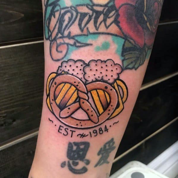 Beer and pretzel tattoo with foam-topped mugs and "EST 1984" text on forearm, featuring bold black outlines and shading.