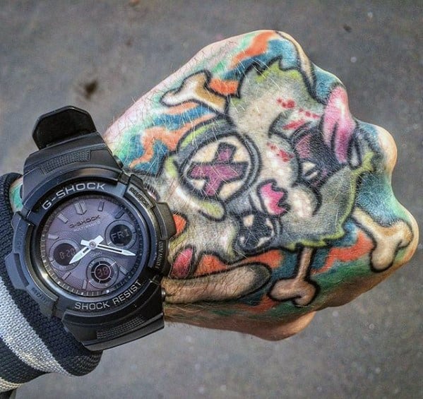 Tattoo of a cartoon skull with pink X eyes, crossbones, and vivid colors on a hand wearing a black G-Shock watch.