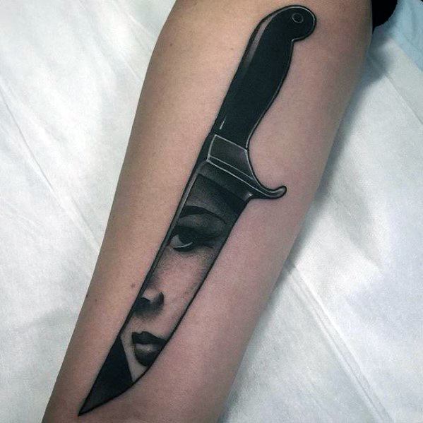 Knife tattoo with a woman's face reflected in the blade, creating a striking illusion of depth and contrast on the forearm.