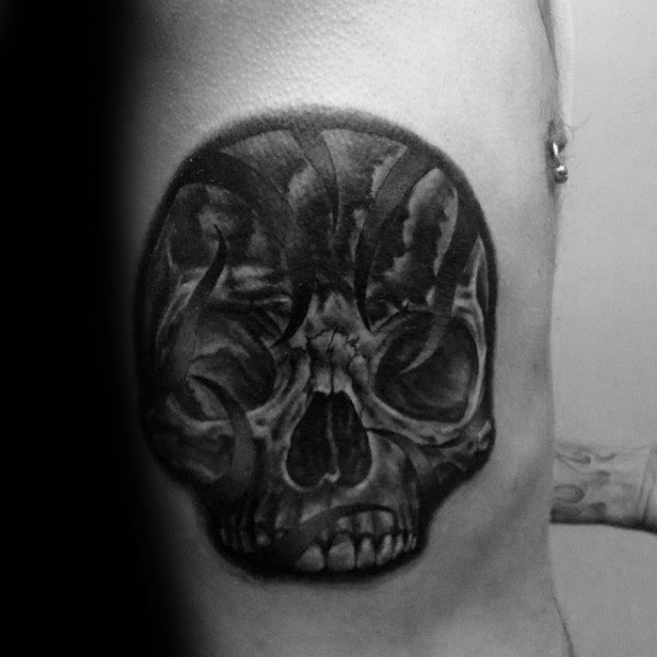 Dark tribal skull tattoo on a man's side, featuring bold black swirls and shading for a dramatic, intense effect.