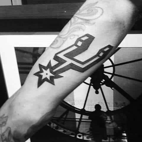 Forearm tattoo of the San Antonio Spurs logo in bold black ink, featuring the signature spur shape with a star in the center.