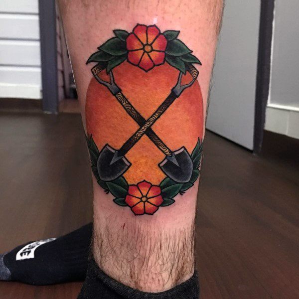 Shovel tattoo design featuring two crossed shovels with intricate handles, framed by bold orange shading, flowers, and green leaves.