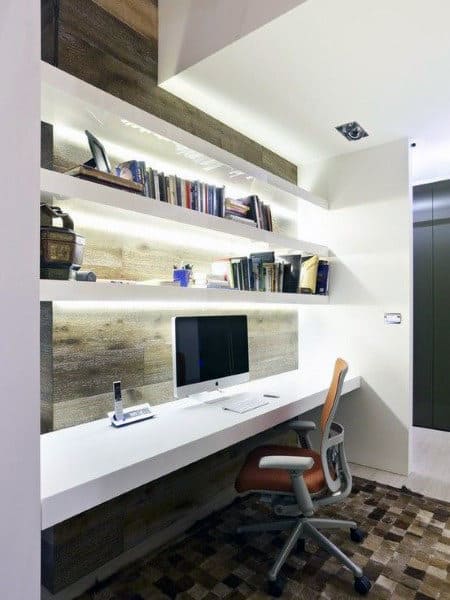 Modern home office with a desk, computer, ergonomic chair, and illuminated bookshelves on a wood-accented wall