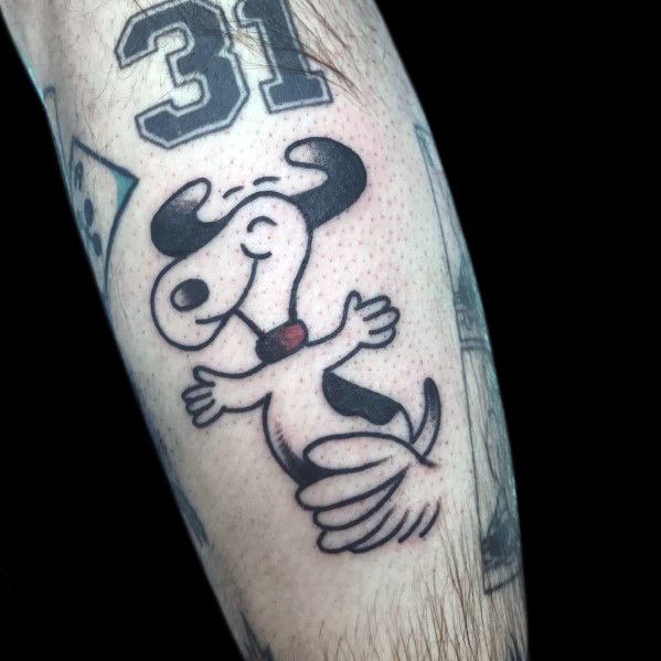 Black and white tattoo of Snoopy joyfully dancing with a red collar, placed on a person's arm among other tattoos.