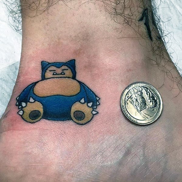 Small Snorlax tattoo on ankle beside a coin for scale, showcasing bold colors and clean outline on lightly hairy skin.