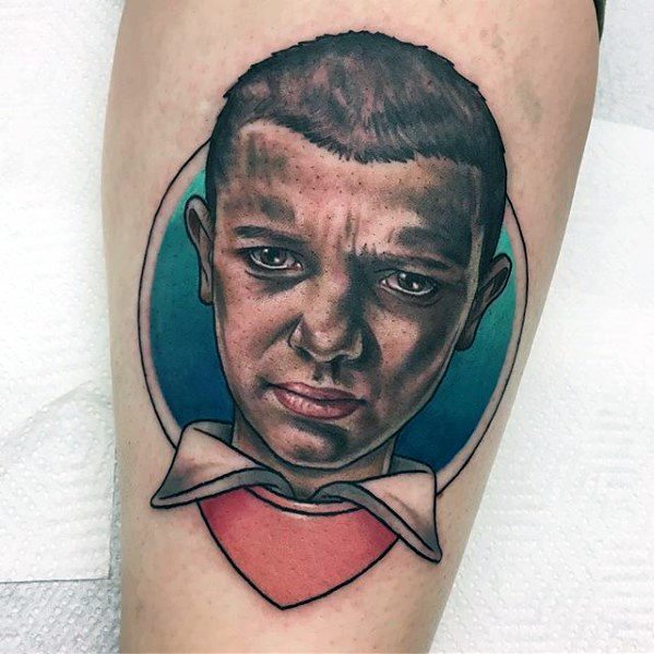 Realistic tattoo of Eleven from Stranger Things on a man's leg, with a serious expression and a bold colored background.
