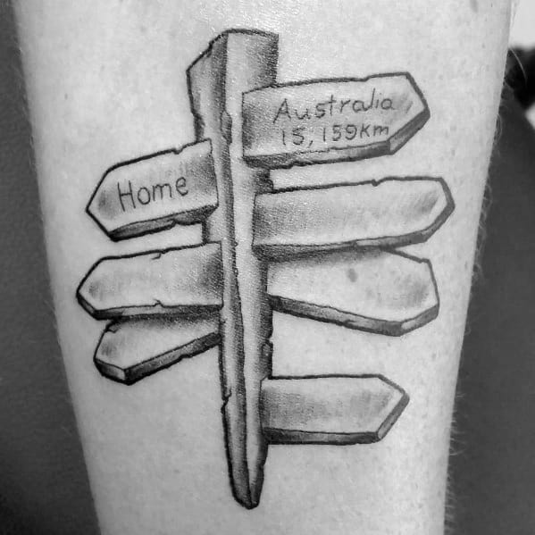 Black and grey tattoo of a weathered street sign with arrows pointing in different directions, including 'Home' and 'Australia 15,159 km.'