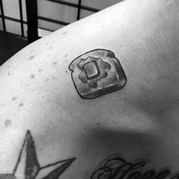 Simple toast tattoo with a square butter pat in the center, inked on the upper arm in black and grey shading for a minimalist look.