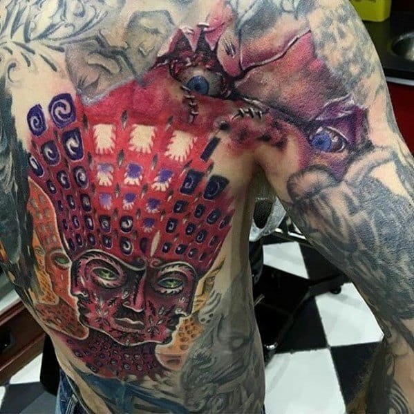 Tool-inspired tattoo on a man's torso featuring a detailed red face, geometric patterns, and realistic eyes on the upper chest.