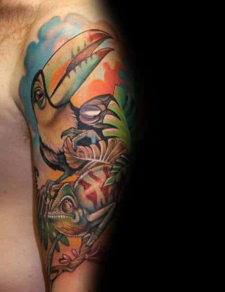 Colorful toucan and chameleon tattoo on upper arm with tropical leaves and vibrant sunset sky in the background.