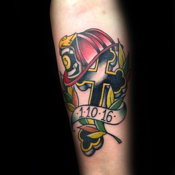 Traditional-style cross tattoo with a firefighter helmet, laurel leaves, and a banner showing the date 1·10·16, inked on the forearm.