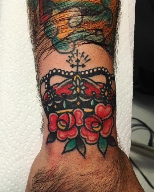 Traditional crown tattoo with bold black outlines, red roses, and jewel accents on a man's wrist, showcasing classic style.