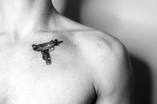 Black ink Uzi tattoo on a man's upper chest, positioned near the collarbone. Bold and detailed firearm design.