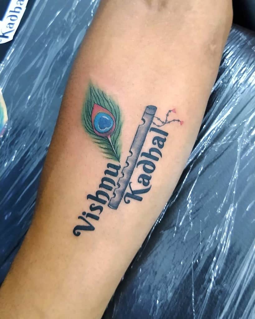 Forearm tattoo featuring a peacock feather, a flute, and the words 'Vishnu Kadhal' in bold script, with fine shading and vibrant colors.