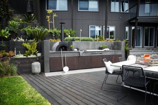 Modern outdoor kitchen with a pizza oven and seating area on a wooden deck, surrounded by lush outdoor greenery
