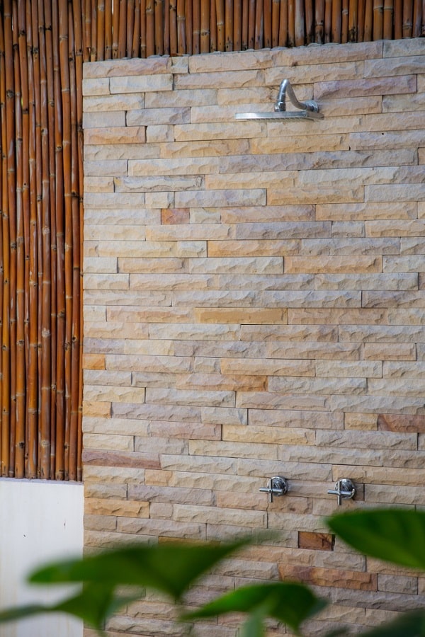 Outdoor shower with stone wall and bamboo paneling surrounded by green plants.