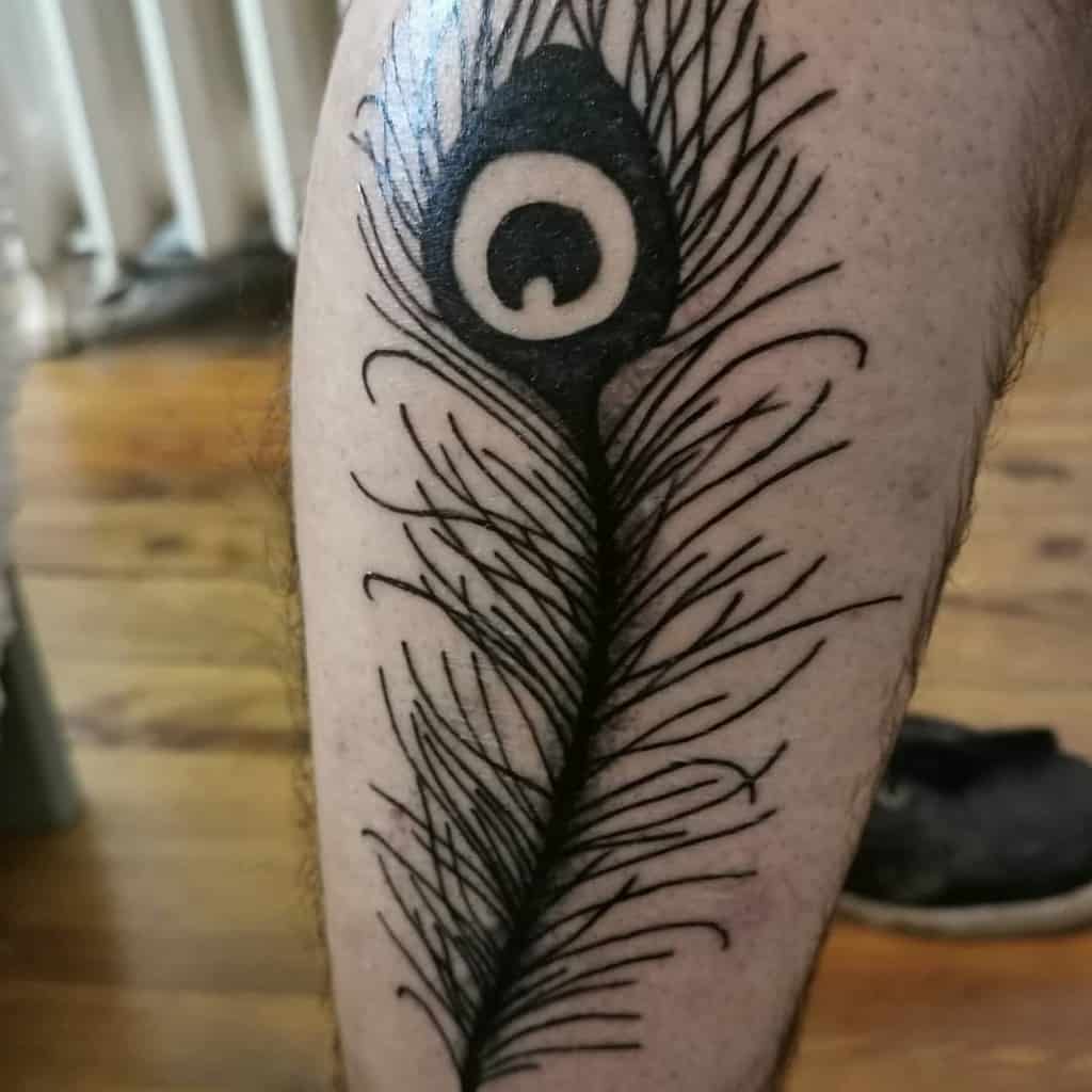 Black ink peacock feather tattoo on the leg, featuring bold linework and a stylized eye pattern at the top with intricate detailing.