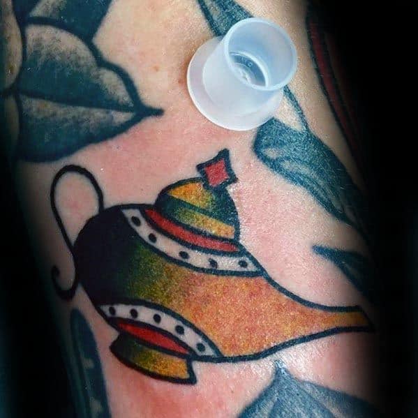 Genie lamp tattoo on skin with a colorful glow, plastic cap resting nearby