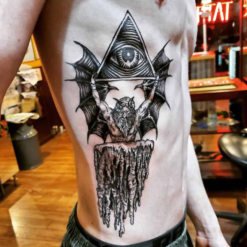 Tattoo of a winged demonic figure holding an all-seeing eye inside a triangle, inked in black and grey shading on the ribcage.