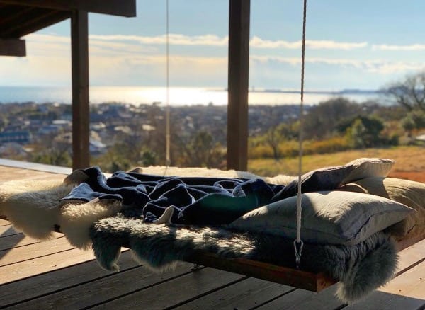 Outdoor hanging bed with cushions and blankets overlooking a scenic view of the sea and landscape.