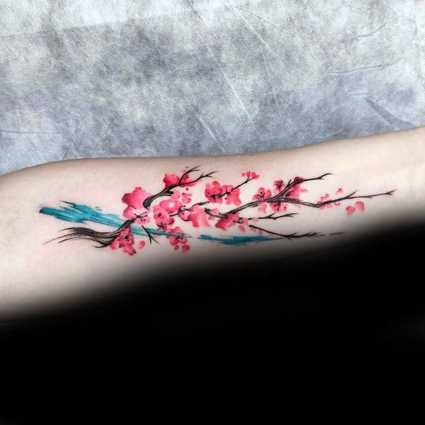 Forearm tattoo with a watercolor-style cherry blossom branch, vibrant pink flowers, delicate black branches, and a blue brushstroke accent