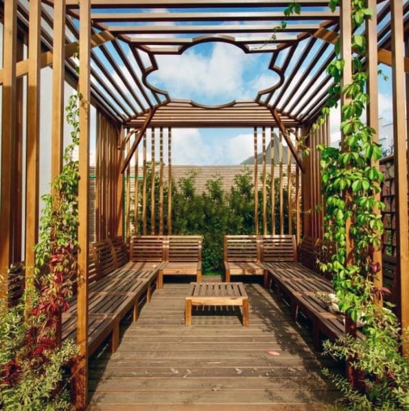 Wooden pergola with intricate ceiling design, built-in benches, and lush climbing plants.