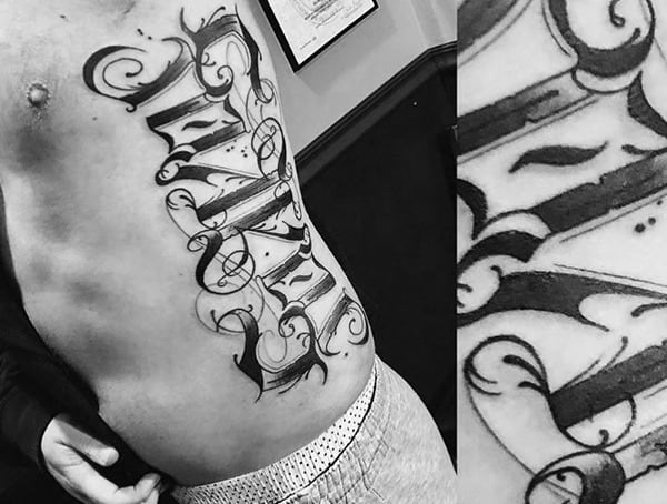Black and grey script tattoo on the ribcage featuring bold, ornate lettering with intricate shading and flourishes in a calligraphic style.