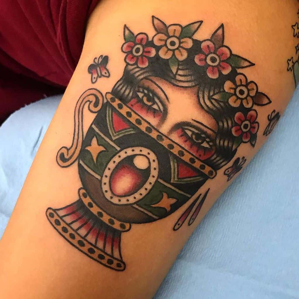 Traditional tattoo of a gypsy woman’s eyes peeking from an ornate goblet, adorned with flowers and bold colors on the arm.