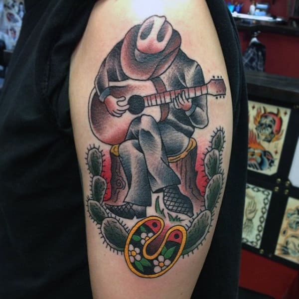 American Traditional Style Tattoo Of Western Man In Desert Playing Guitar On Mans Bicep