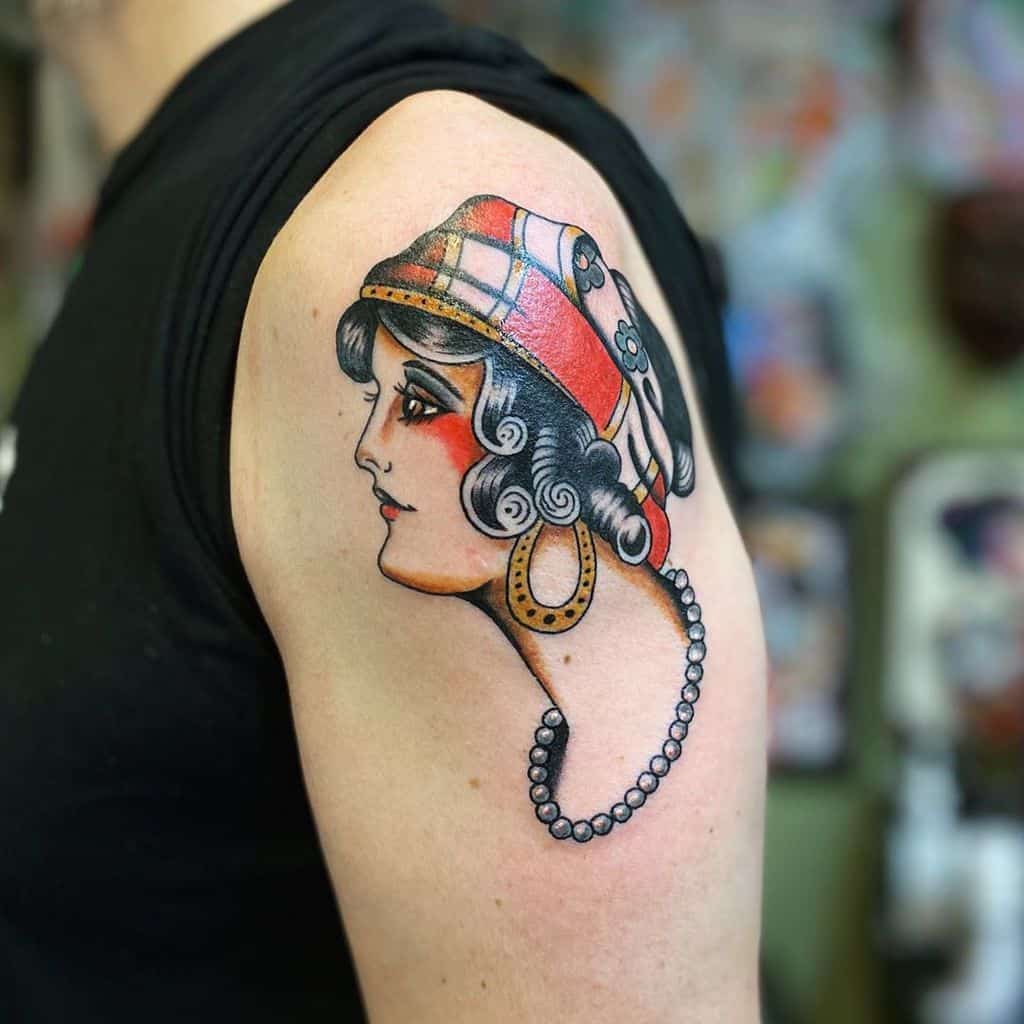 Traditional tattoo of a gypsy woman in profile with a plaid headscarf, gold earrings, and a pearl necklace on the upper arm.