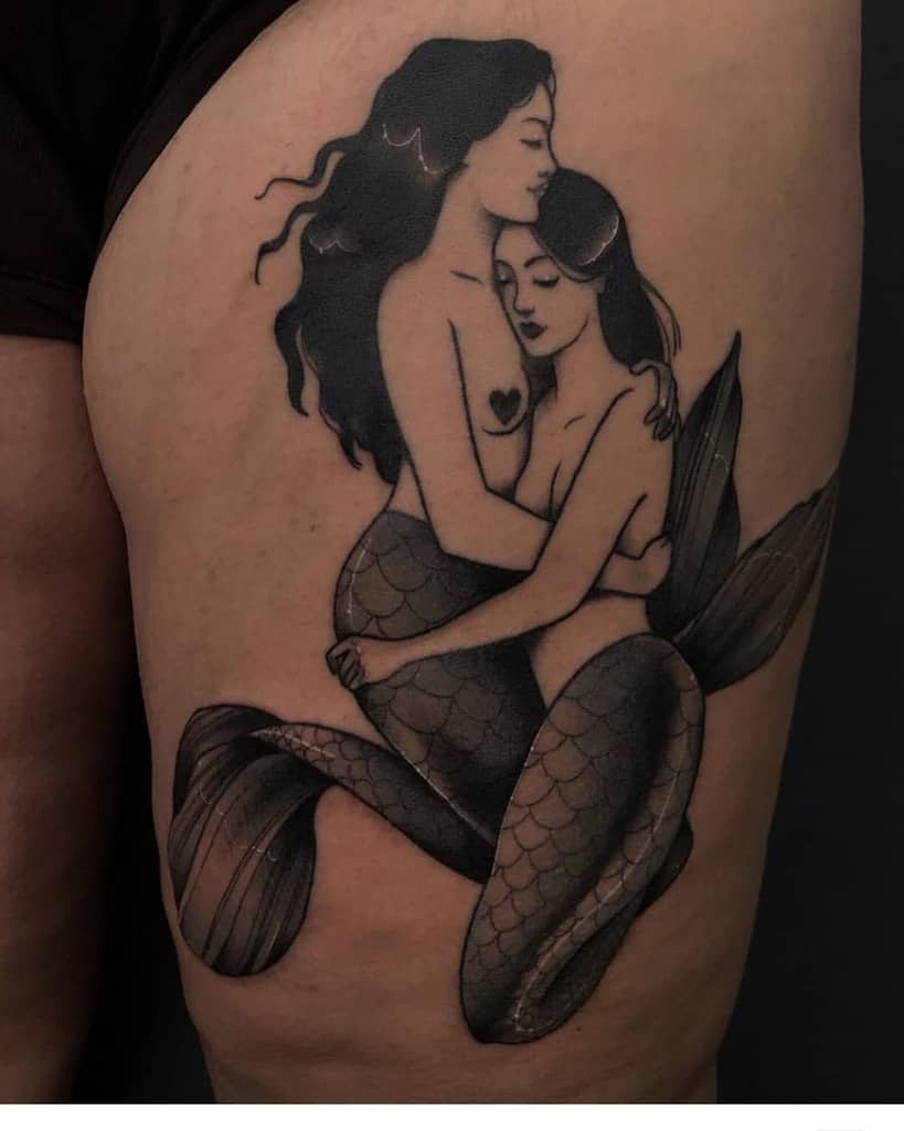 Black and grey tattoo of two mermaids embracing lovingly, with detailed scales and flowing hair, inked on the upper thigh.
