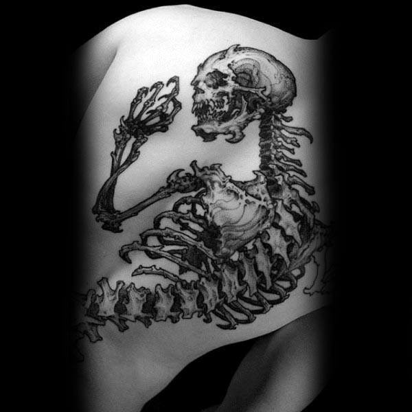 Detailed black ink tattoo of a full human skeleton in a curled position, spanning the shoulder blade and upper back.