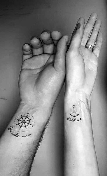 Matching wrist tattoos of a compass with "Guide me" and an anchor with "Hold me," symbolizing direction and stability in love.