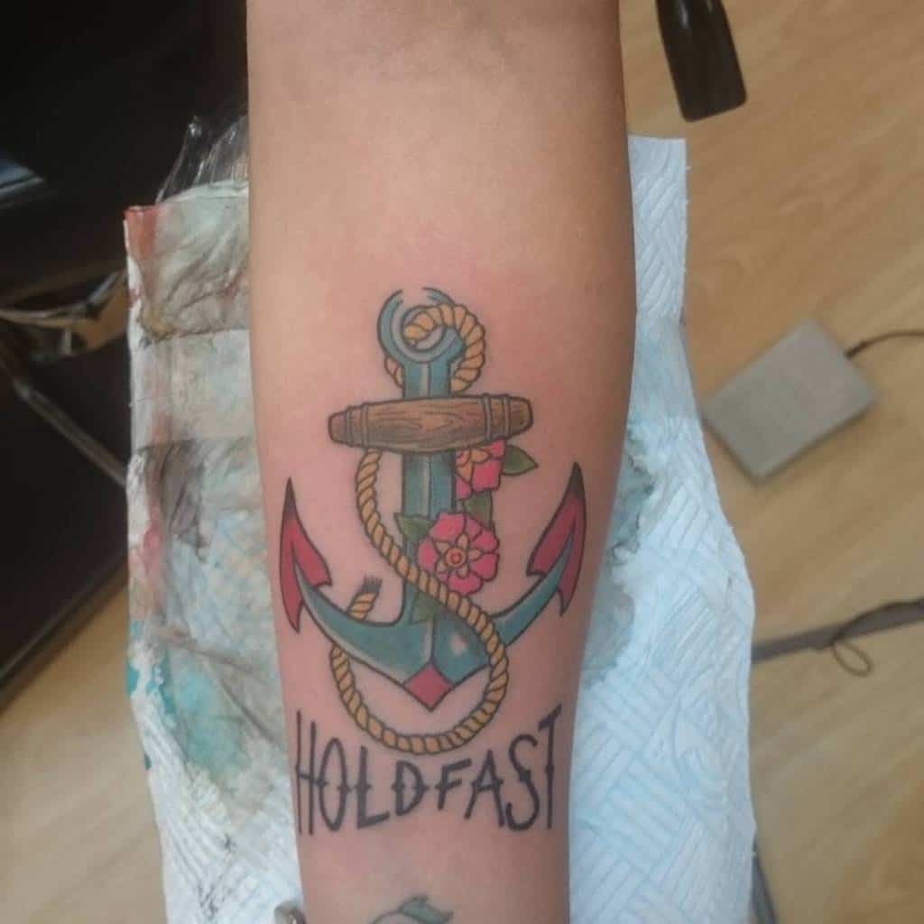 Tattoo of an anchor with flowers and rope, reading "HOLD FAST" on forearm