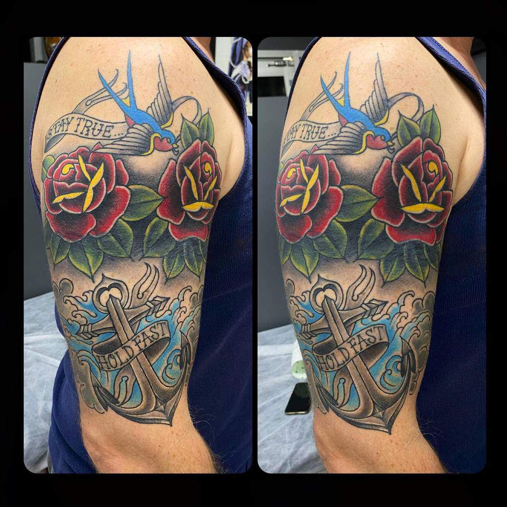 Colorful tattoo of a blue bird, roses, and an anchor with "Hold Fast" on an upper arm
