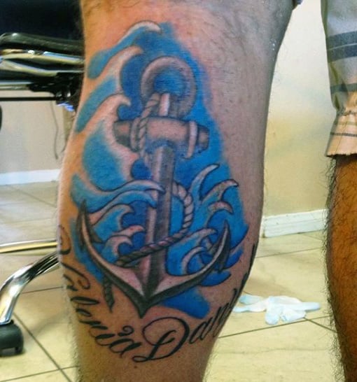 Tattoo of an anchor with blue waves and cursive text on a calf