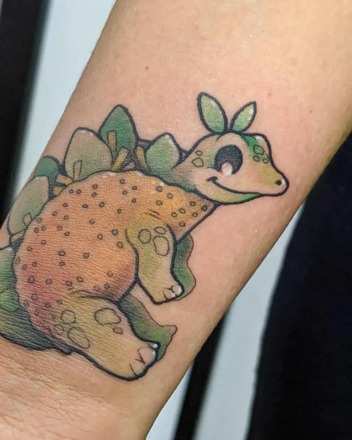 Tattoo of a cute, colorful dinosaur with a big smile, green leaves on its back, and dots on its body