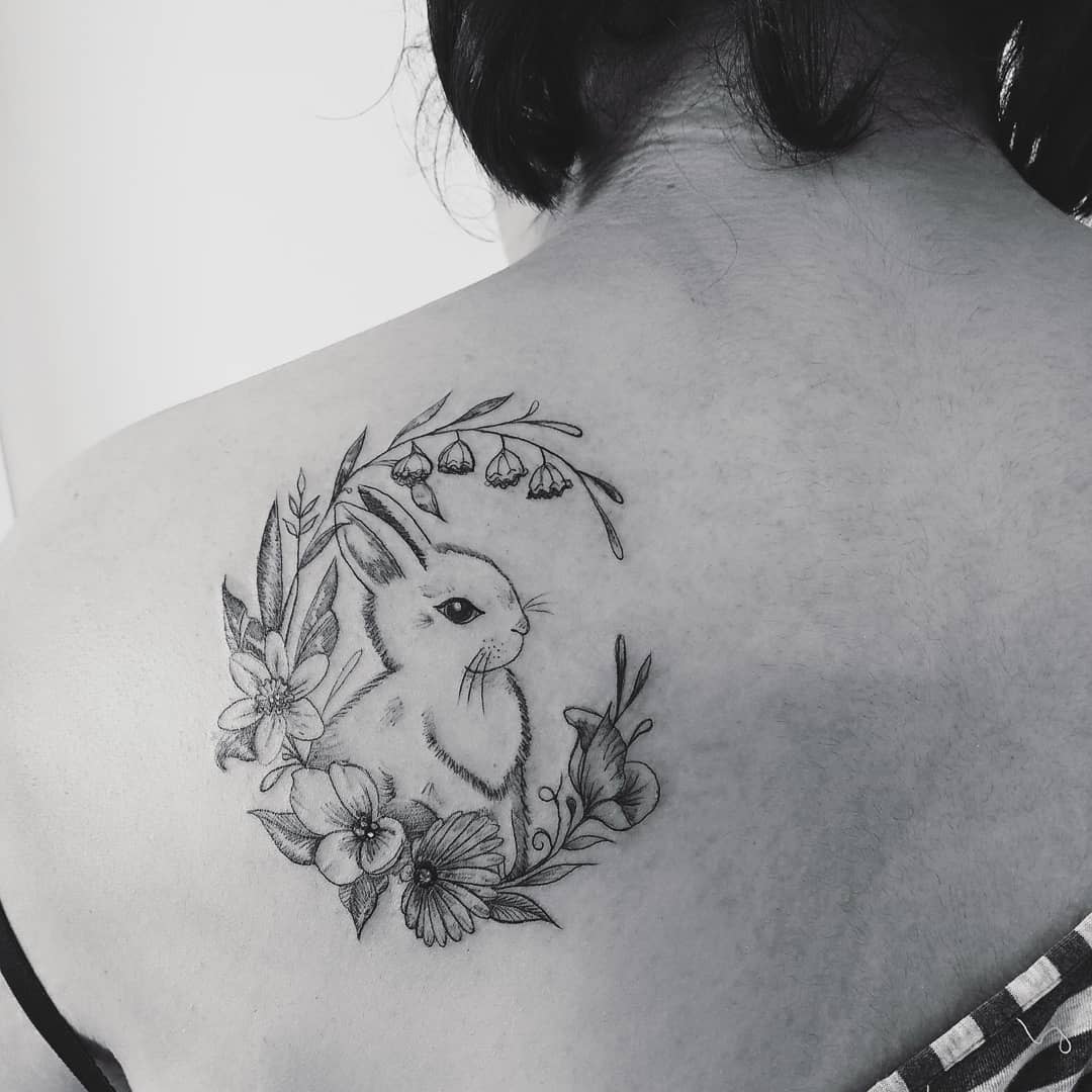 Delicate black and grey tattoo featuring a rabbit surrounded by a floral wreath, including flowers and leaves