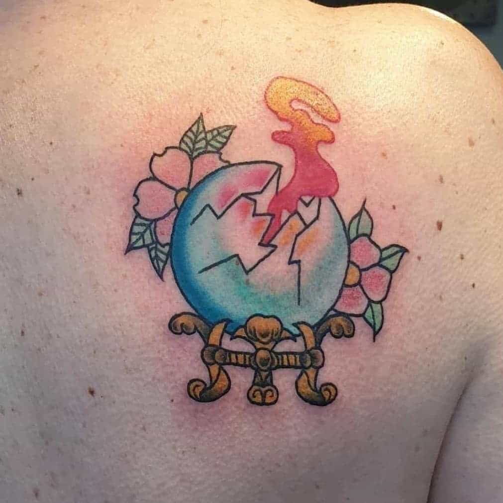 Tattoo of a cracked egg with colorful smoke, surrounded by flowers, and standing on golden stand
