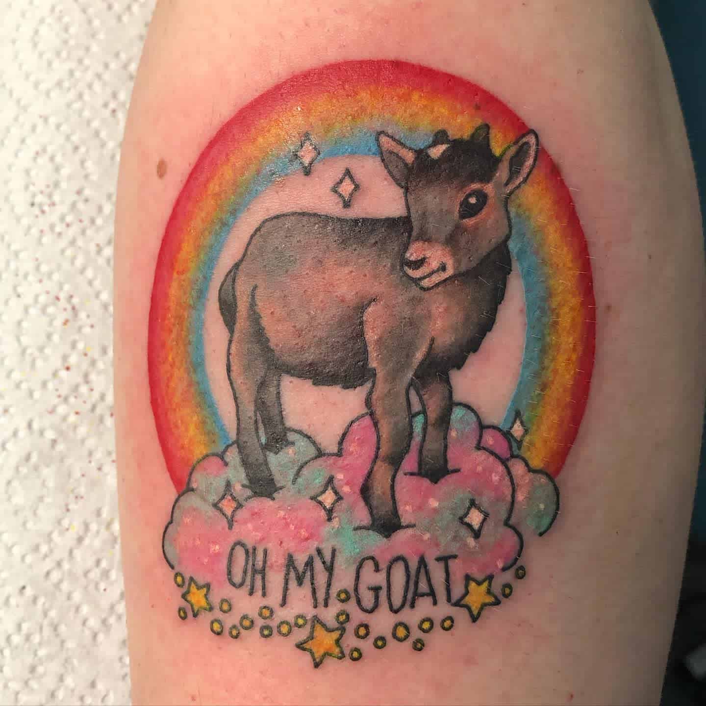 Tattoo of a goat standing on clouds with a rainbow background, surrounded by stars and sparkles, with "Oh My Goat" text below