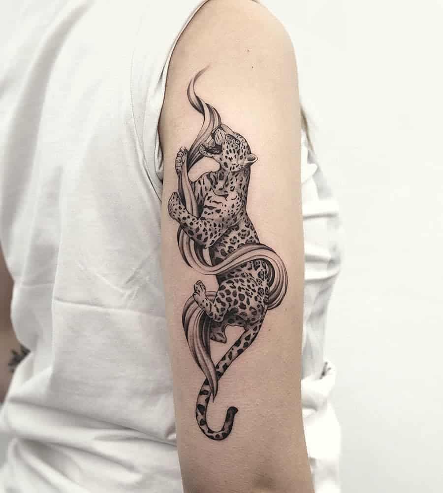 A graceful black and grey tattoo of a jaguar playfully interacting with flowing ribbons, wrapping around the upper arm with smooth, dynamic lines.