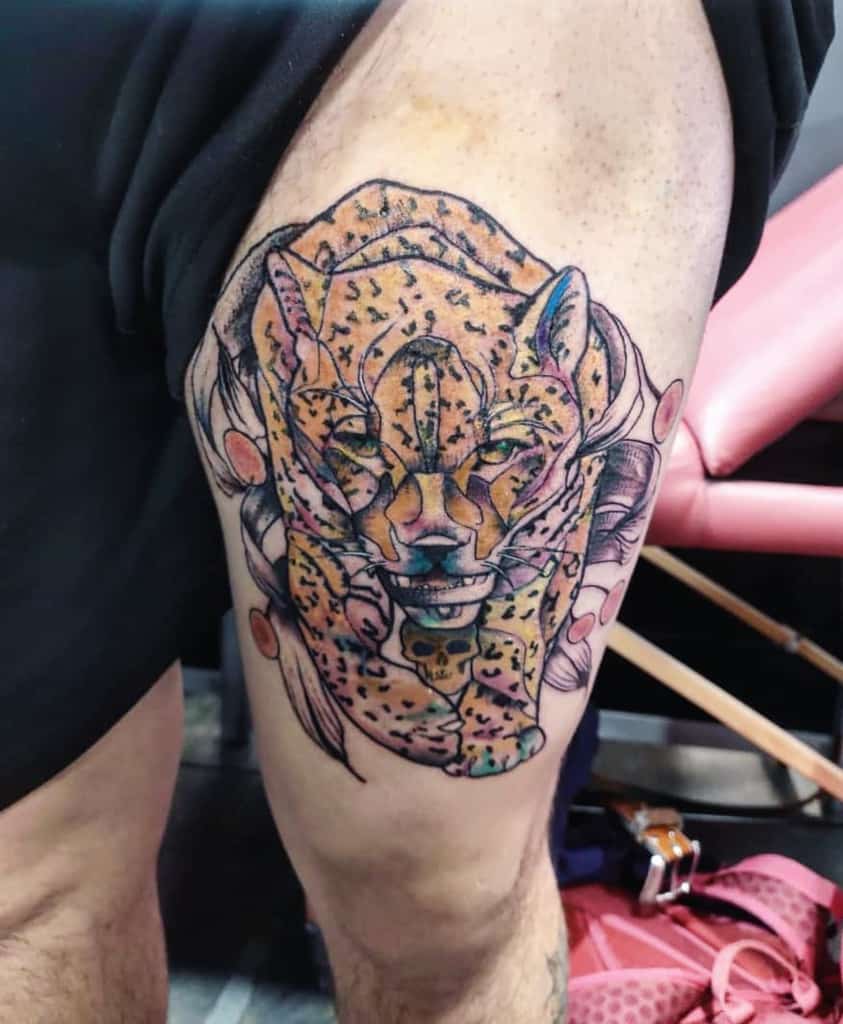 A colorful tattoo of a jaguar's face surrounded by delicate flowers, with the jaguar appearing to be wrapped in the floral elements, placed on the upper thigh.