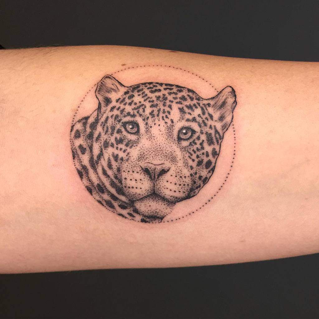 A finely detailed black and grey tattoo of a jaguar's face, framed within a dotted circular design, placed on the forearm for a clean and elegant look.