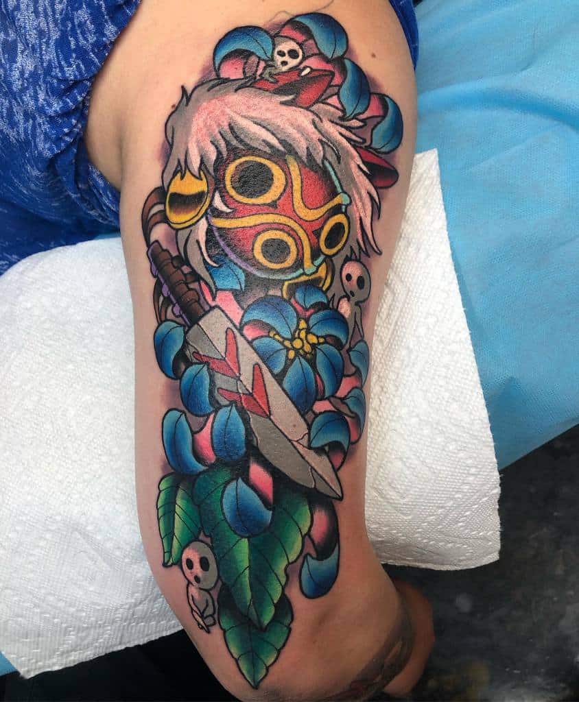 Colorful tattoo of a character wearing a wolf mask, holding a sword, surrounded by blue flowers and small skull details. The design features vibrant shading and intricate leaf patterns, creating a bold and lively scene on the upper arm.