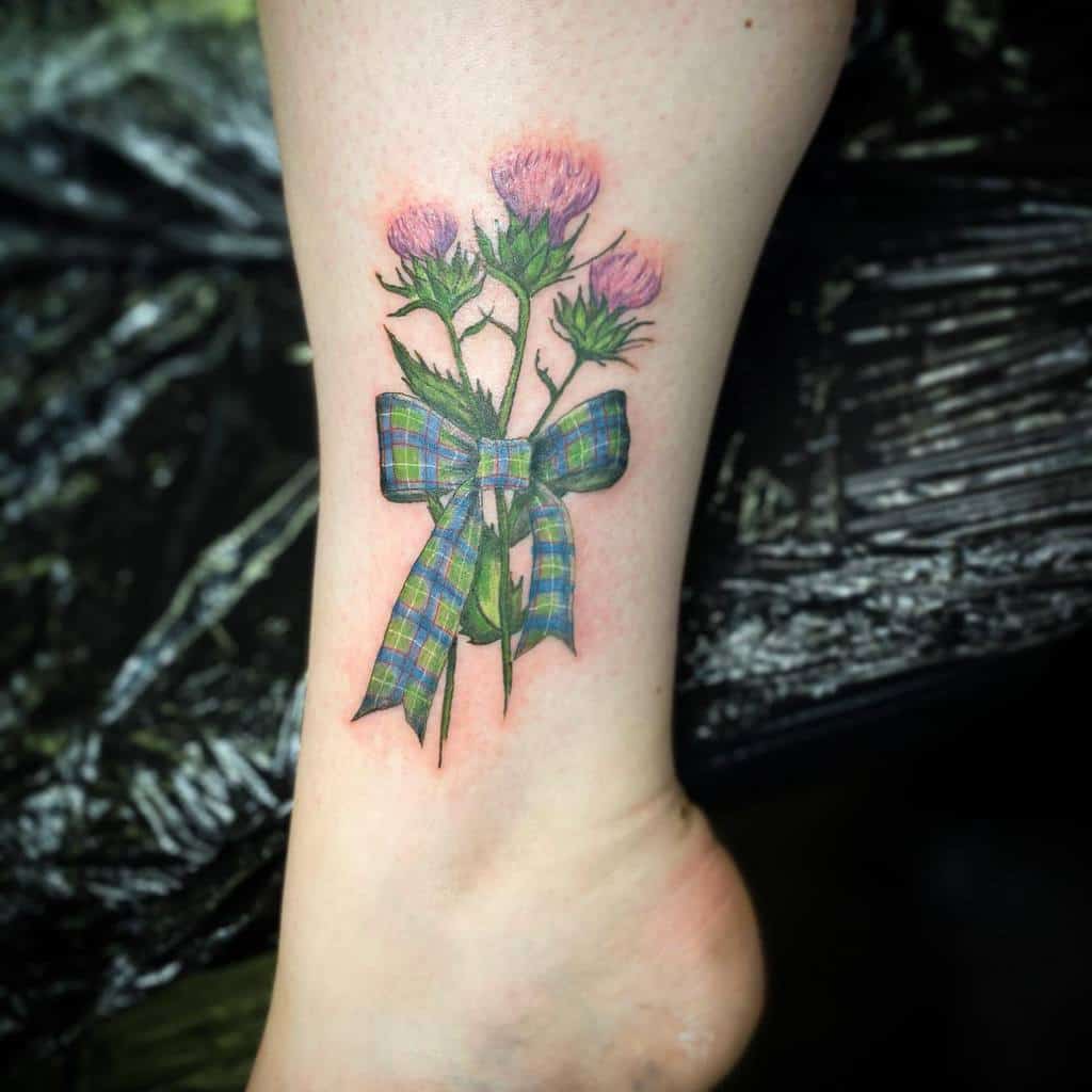 A tattoo featuring delicate thistle flowers gracefully entwined with a blue plaid ribbon adorns a lower leg