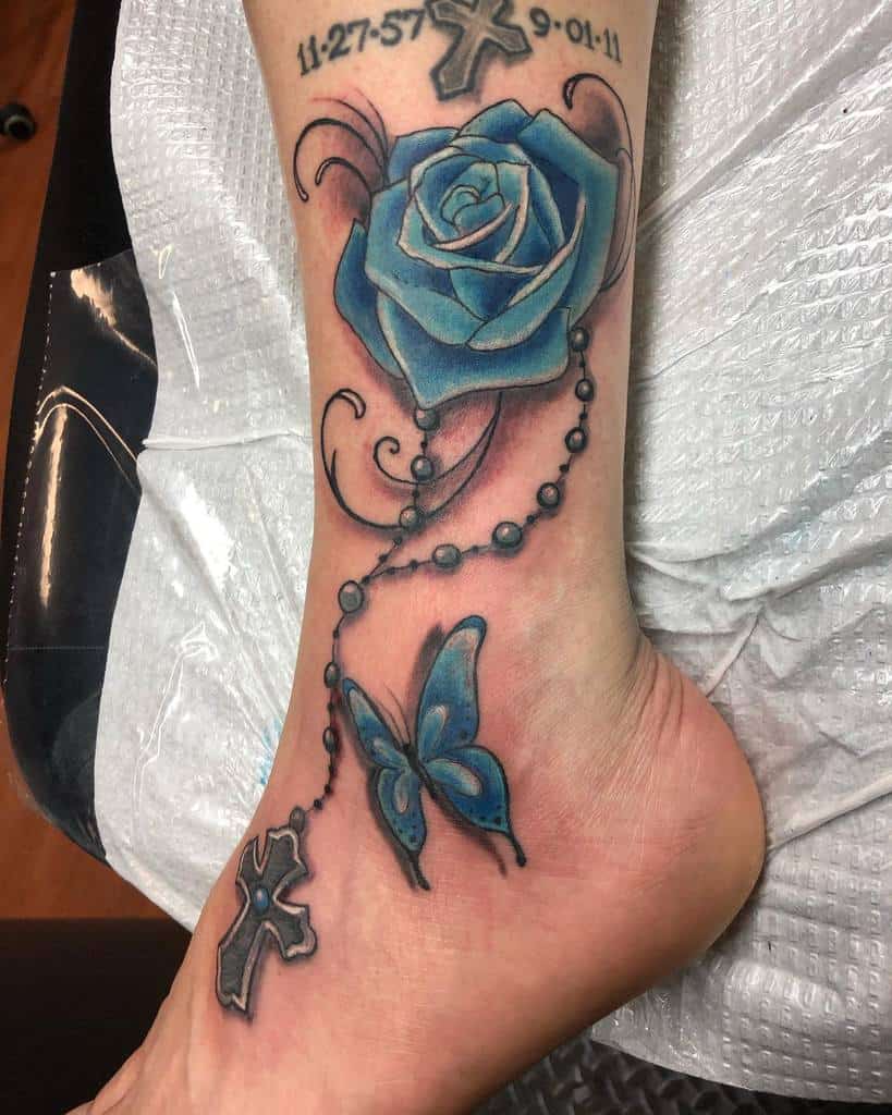A stunning blue rose tattoo graces the ankle, intertwined with delicate butterflies, ornate crosses, and an elegant beaded chain design