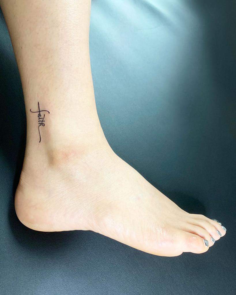 A minimalist ankle tattoo featuring the word "faith" elegantly intertwines with a subtle cross on a foot
